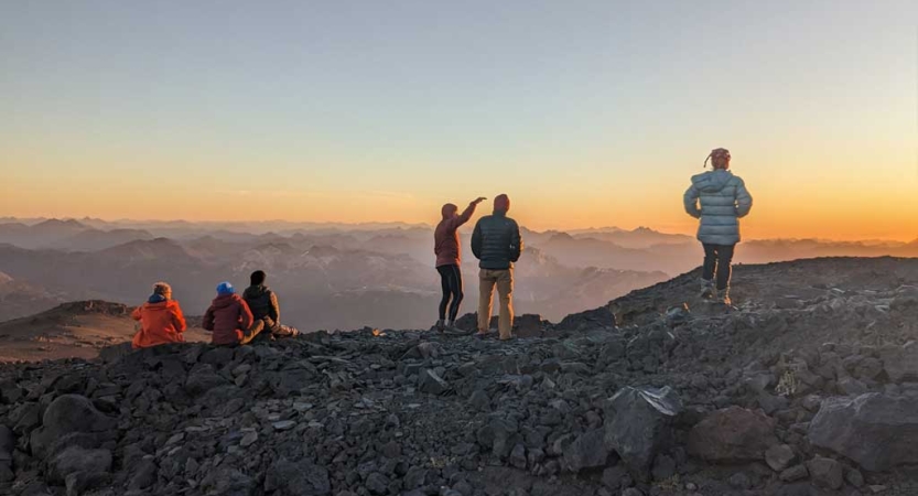 A group of people stand on a rocky overlook, taking in the vast mountain view. The sky appears in soft colors, as the sun is either rising or setting. 